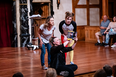 ASC-Complete_Works-Cast Program-09934 Jenny Bennett sitting on the front of the stage and wearing black shirt and pants holds up the play program for Ginna Hoben in white t-shirt and geans, and Allie Babich in black and gray t-shirt ad red and yellow tights, both leaning over to read what Bennett is pointing at in the program